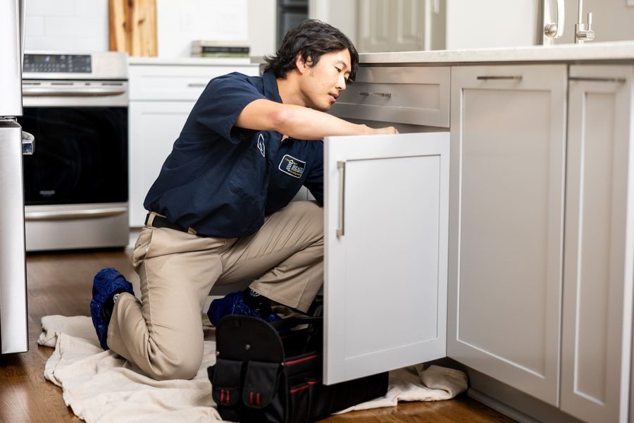 Repair technician kneels on a cloth in a kitchen, reaching into an open cabinet with a toolbox nearby.