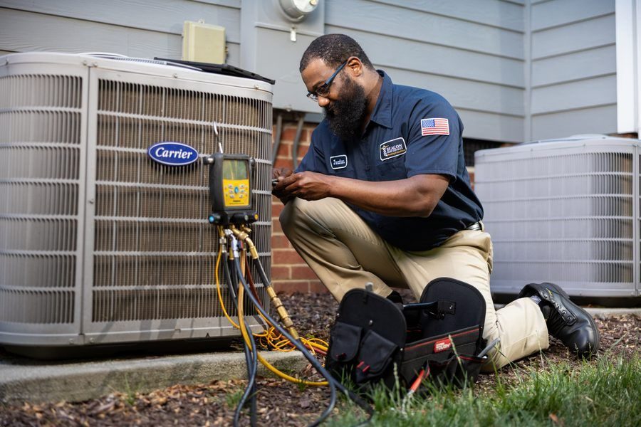 Heating & Air technician kneeling beside a Carrier outdoor unit, testing with diagnostic gauges and wires.